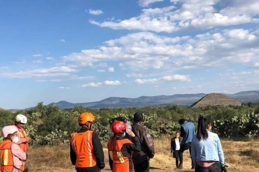 A group of people in orange vests standing in a field with a mountainous landscape in the background. 1
