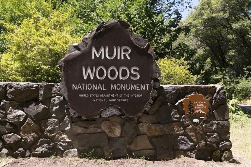 A sign for Muir Woods National Monument with a stone wall backdrop. 1