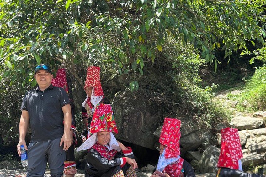A group of people in traditional attire with red head coverings, standing and sitting in a natural setting with greenery and rocks. 4