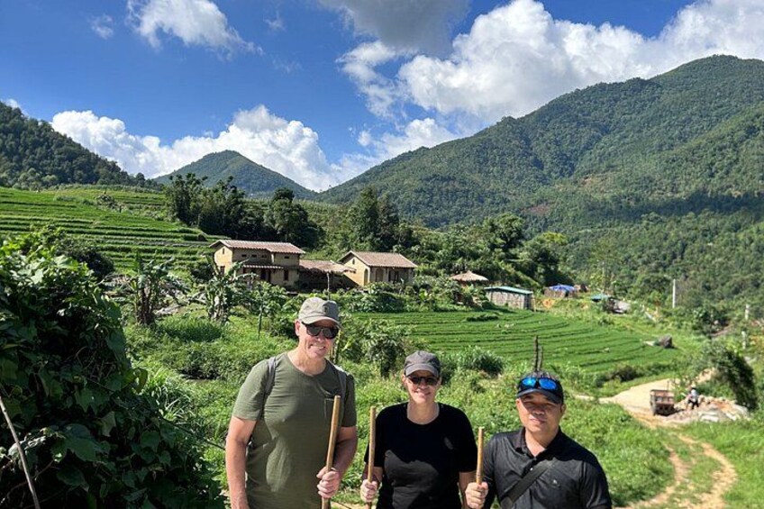 Three people standing in a rural landscape with terraced fields, houses, and mountains in the background. 1