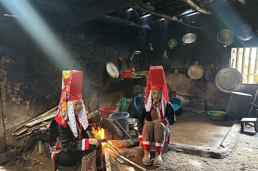 Two individuals in traditional attire with head coverings, a fire burning in the foreground, and various household items in a rustic setting. 2