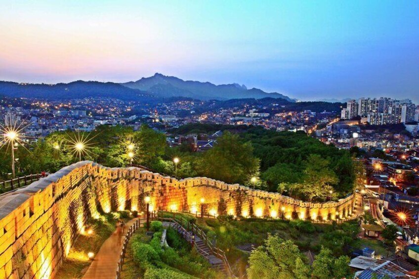 A historic stone wall with a pathway, surrounded by greenery and city lights in the background. 1