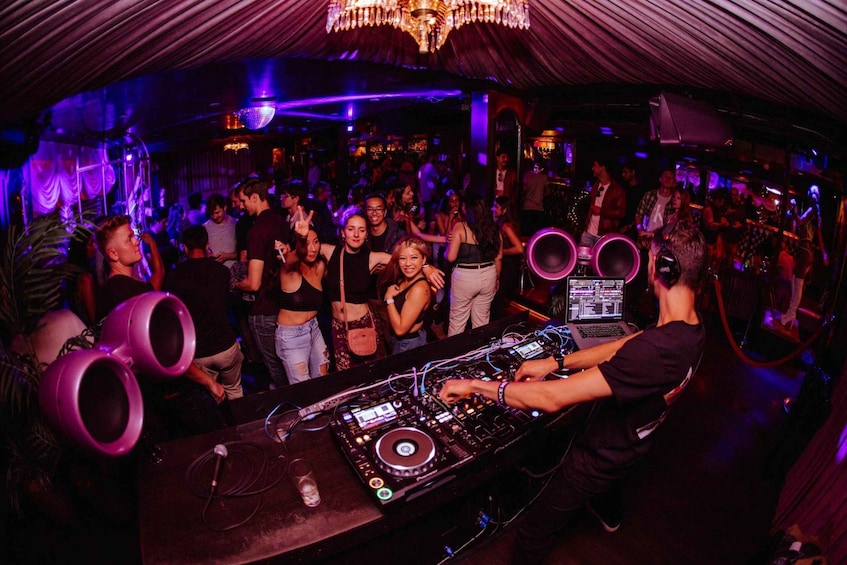 A crowded indoor event with a DJ booth, purple lighting, and a crowd of people. 1