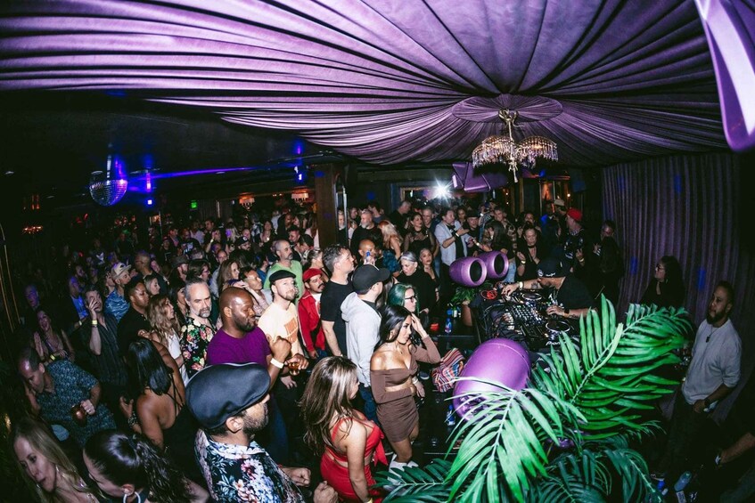 A crowded indoor event with a DJ booth and a purple draped ceiling. 1