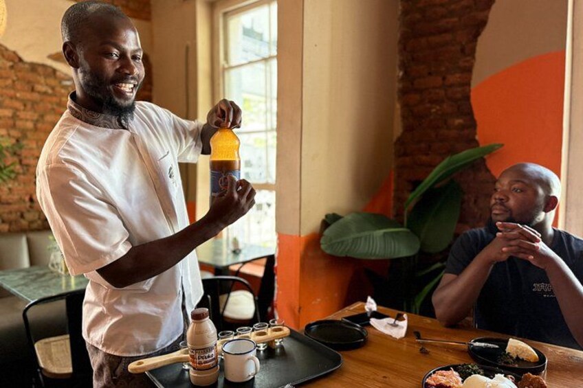 A person pouring a drink into a glass, a table with food, and a plant in the background. 2