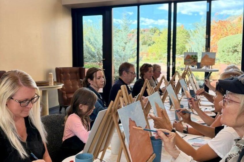 A group of people painting at easels in a well-lit room with large windows. 1