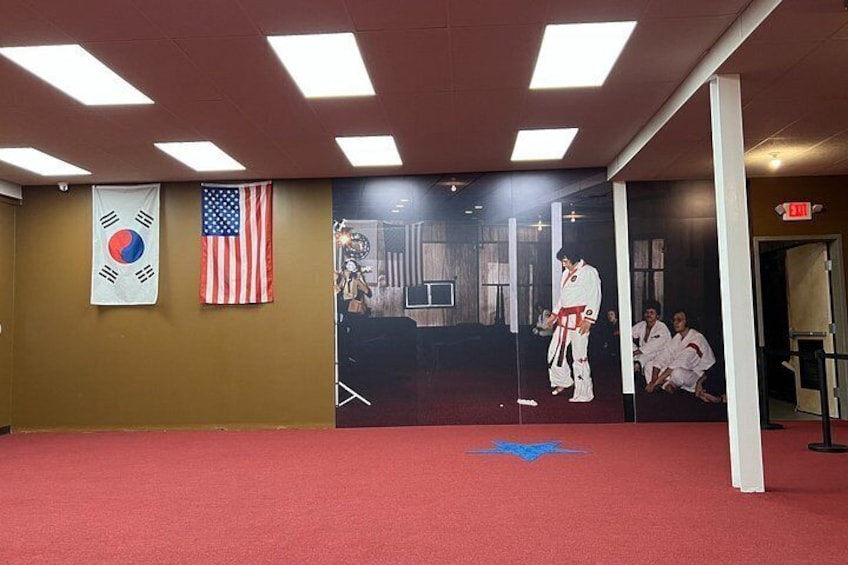 A martial arts training room with a red carpet, a Korean flag, an American flag, and a poster of a martial artist. 2