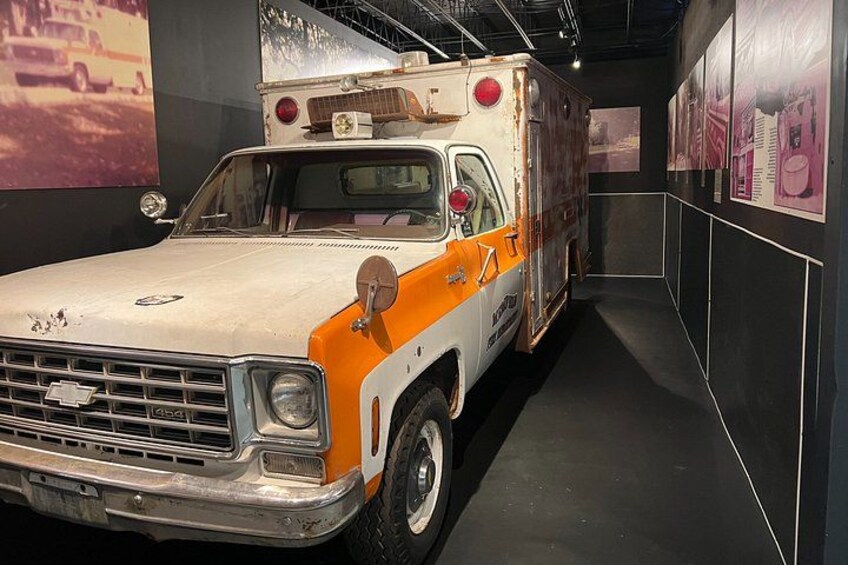 A vintage ambulance with orange and white paint, parked in a narrow indoor space with walls adorned with posters. 3