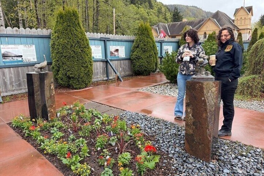 Two people standing in a garden with a wooden fence, a stone monument, and a building in the background. 4
