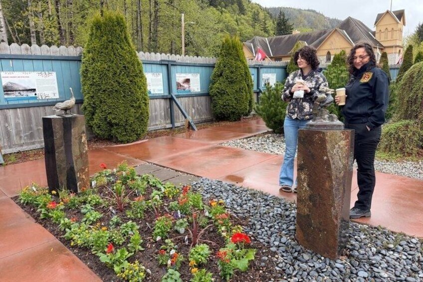 Two people standing in a garden with a wooden fence, a stone monument, and a building in the background. 4