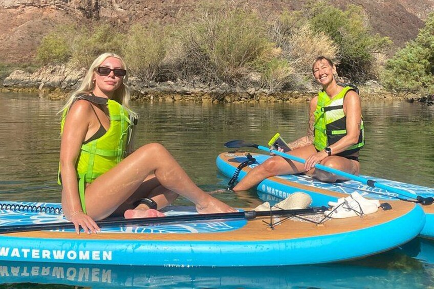 Two people on a blue watercraft with 'WATERWOMEN' written on it, sitting in a river with trees and rocks in the background. 3