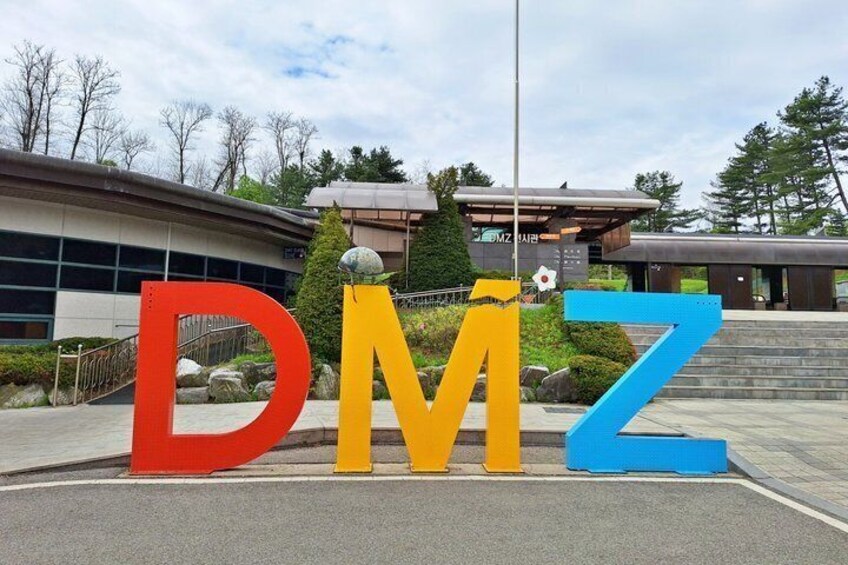 Large letters 'DMZ' in red, yellow, and blue colors, with a building in the background. 1