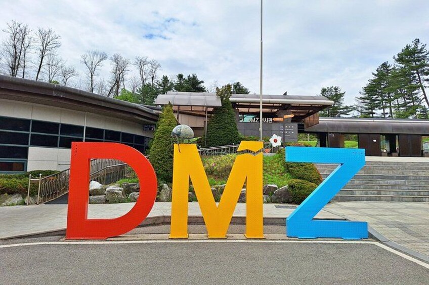 Large letters spelling 'DMZ' in a parking lot with buildings and trees in the background. 1