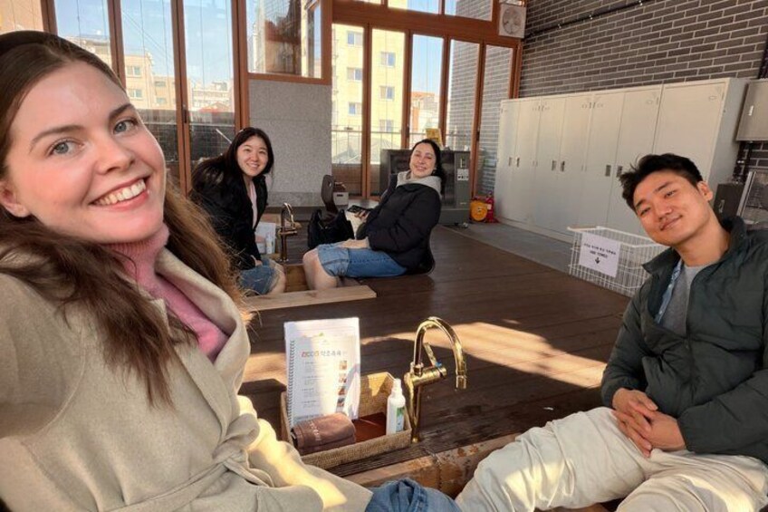 A group of people sitting on a wooden floor in a room with large windows, lockers, and a whiteboard. 1