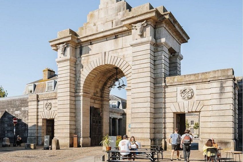 A stone archway with inscriptions, a paved area, and people walking. 2