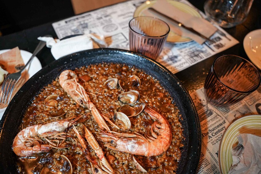 A seafood paella with shrimp and clams in a black pan, set on a table with newspapers and glasses. 1