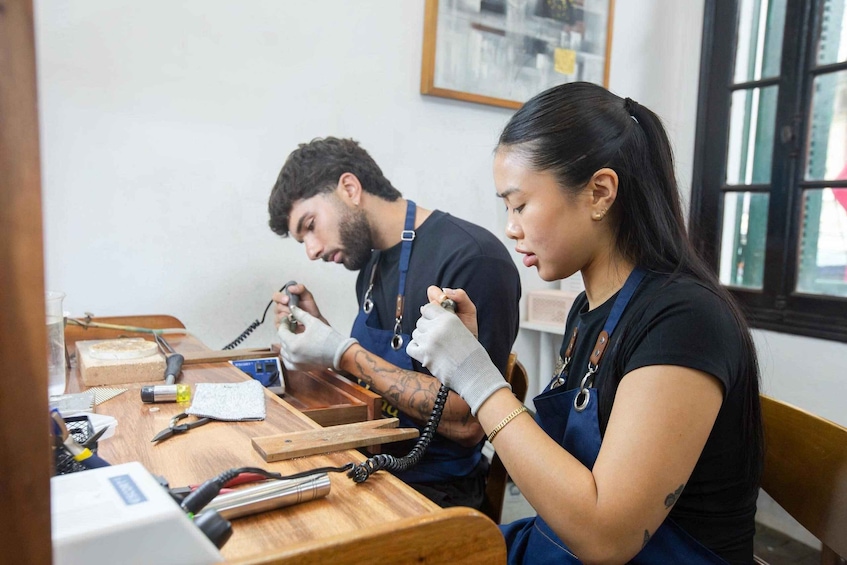 Two people working at a wooden table with tools and a laptop, in a room with a window and framed pictures on the wall. 5