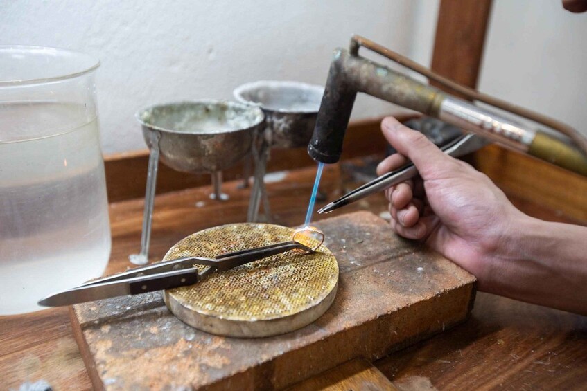 A person working on a metal object with a hammer and a tool, on a wooden workbench with a metal bowl and a glass of water. 2
