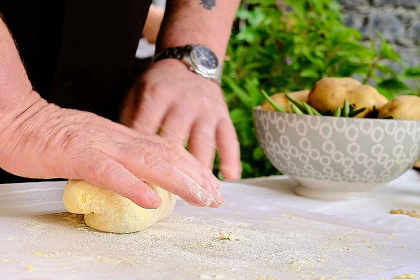 A person kneading dough on a table with a bowl of potatoes in the background. 2