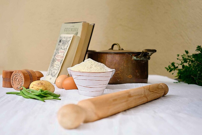 A wooden rolling pin, a bowl of flour, eggs, green beans, a potato, and a book on a table with a pot in the background. 5
