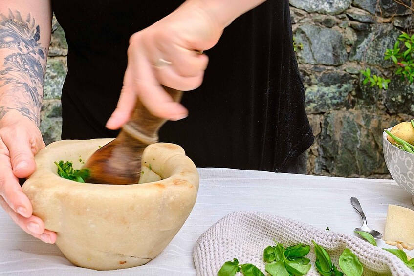 A person using a mortar and pestle outdoors, with a bowl of greens and a cheese block nearby. 1