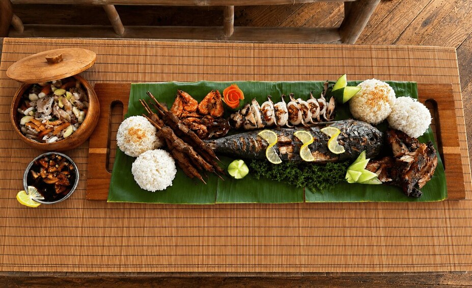 A traditional meal with grilled fish, rice, and vegetables on a banana leaf, served on a wooden table. 3