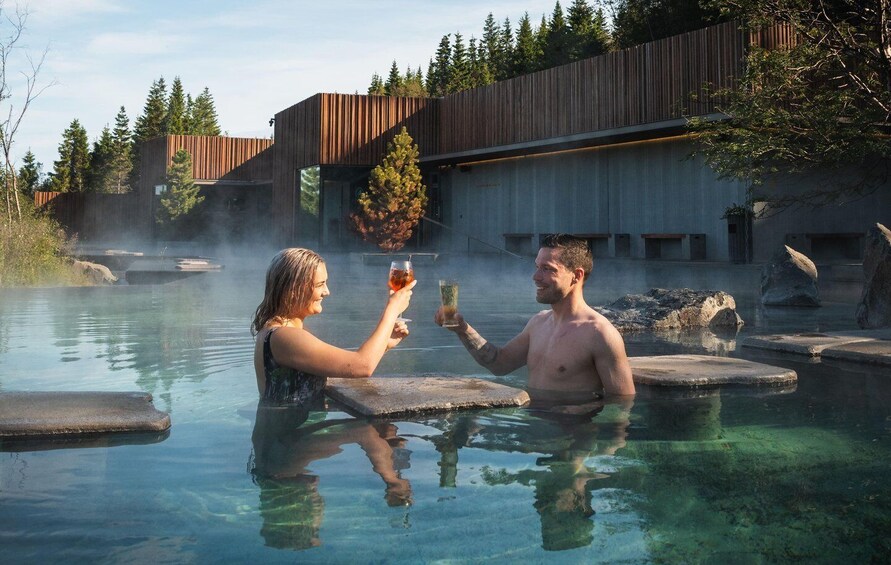 Two people toasting in a clear pool with steam rising, surrounded by wooden structures and trees. 1