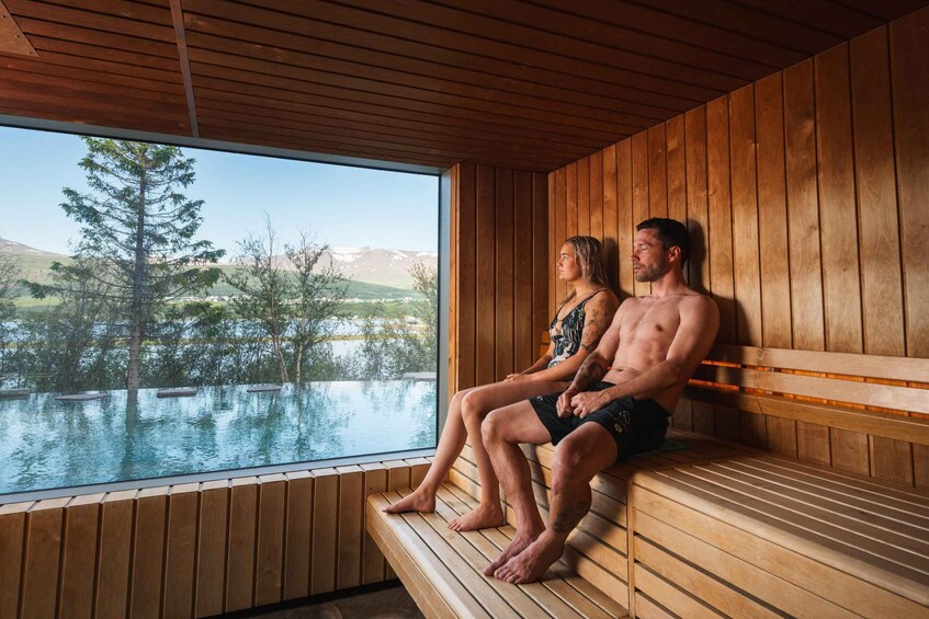 Two people sitting on wooden benches by a large window overlooking a body of water and trees. 3