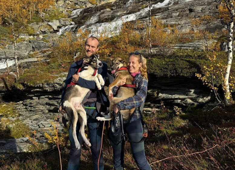 Two people holding dogs in a rocky outdoor setting with autumn foliage. 2