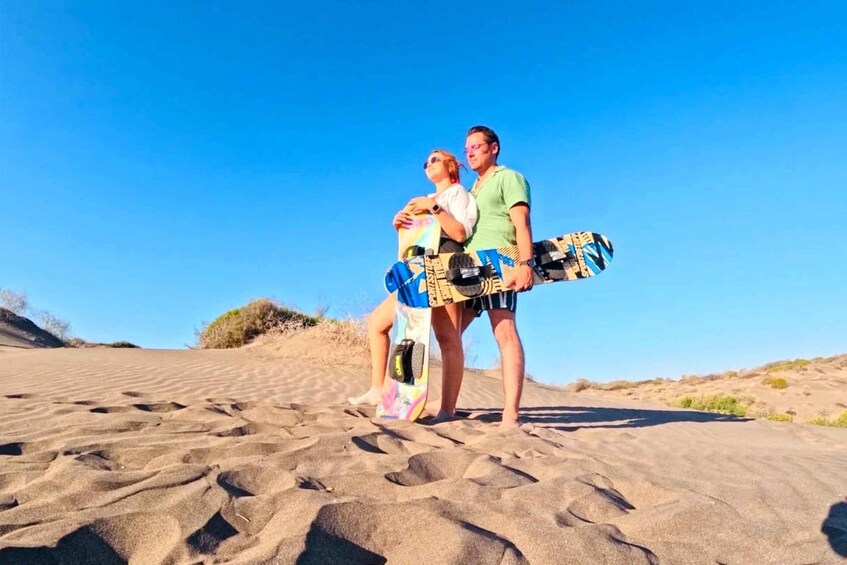 Two people standing on a sandy beach with a clear sky, holding a colorful surfboard. 1