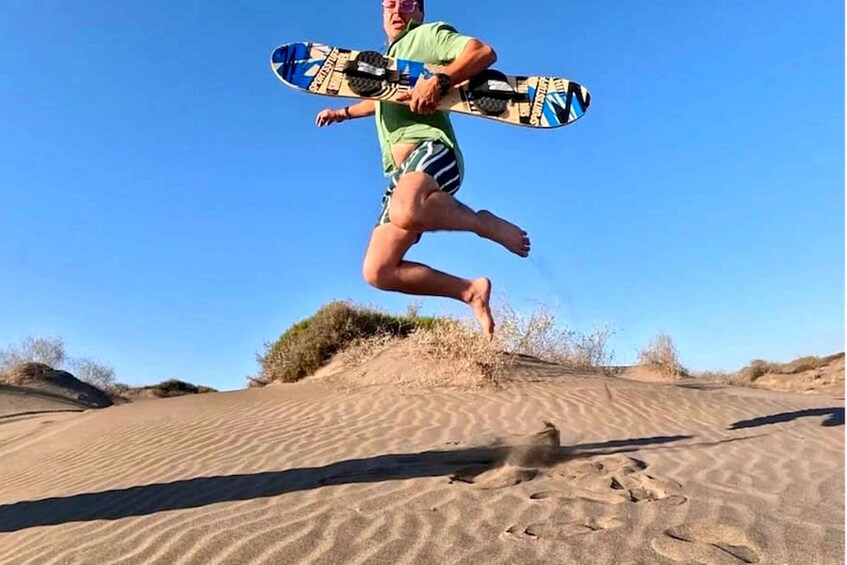 A person mid-jump with a snowboard over a sandy dune landscape. 5