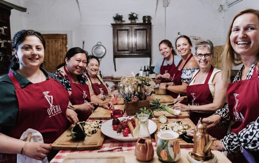 Un gruppo di persone indossa grembiuli rossi con il logo 'OPERA IN THE KITCHEN' e si dedica alla preparazione del cibo attorno a un tavolo di legno. 5