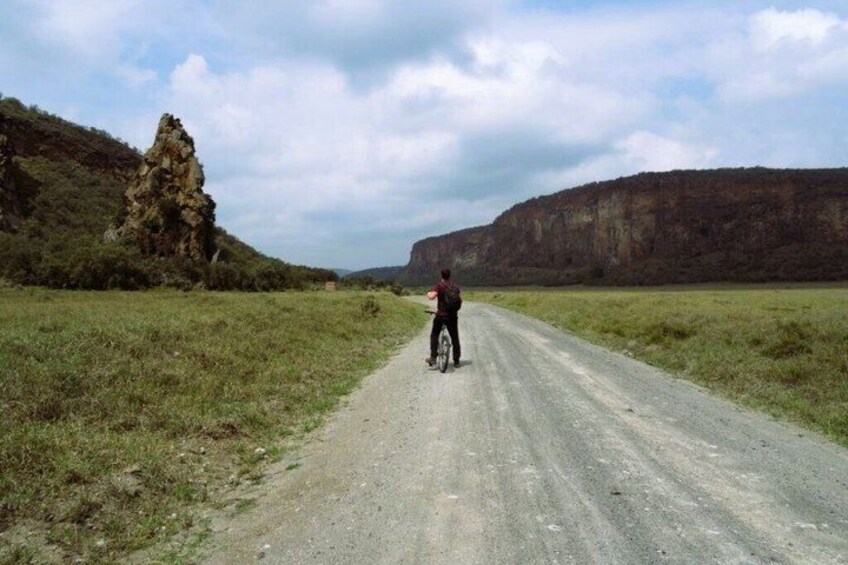 Une personne à vélo sur un chemin de terre, avec des champs verdoyants et des falaises rocheuses en arrière-plan. 2