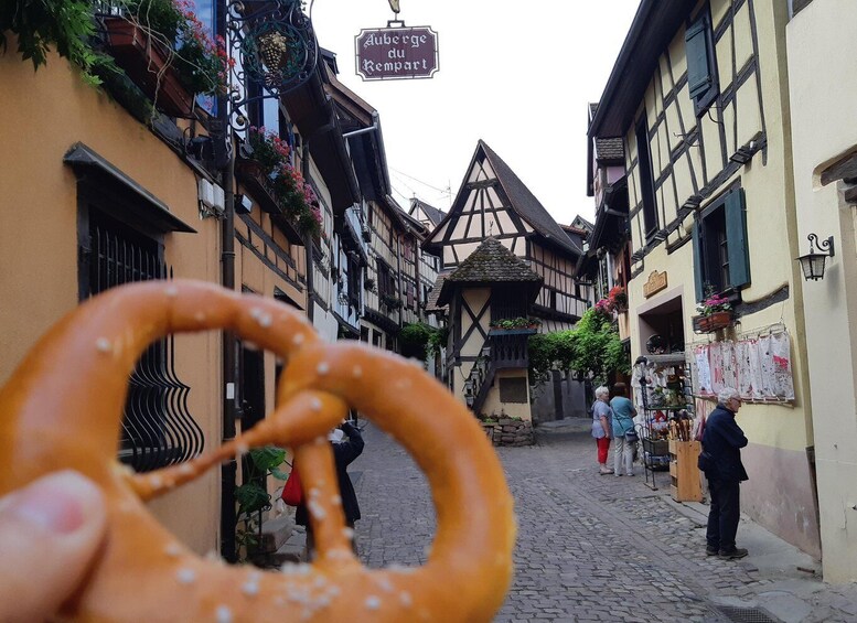 A narrow cobblestone street with traditional half-timbered buildings, a pretzel in the foreground, and a sign reading 'Auberge du Kempart.' 3