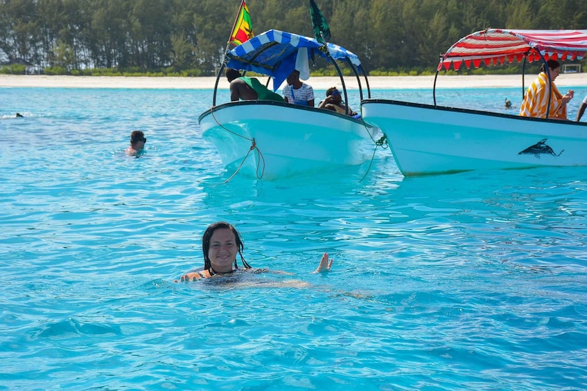 A person swimming in clear blue water with a boat and a beach in the background. 1