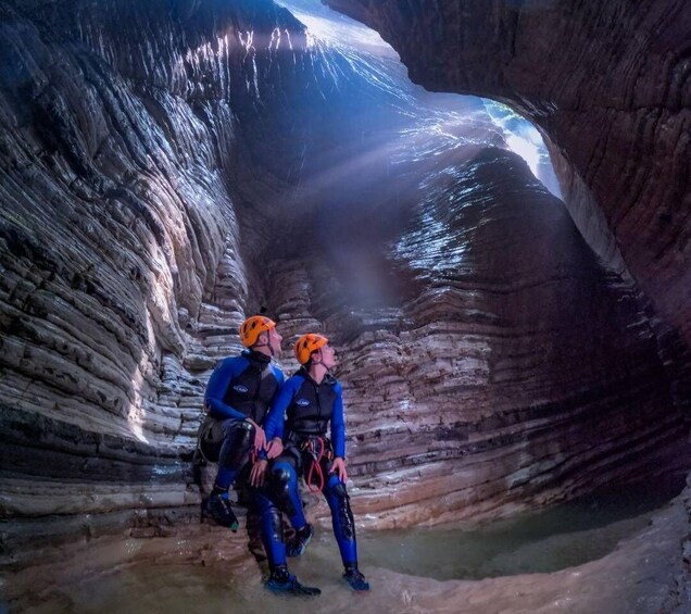 Two people in blue and orange helmets inside a narrow canyon with water at the bottom. 1