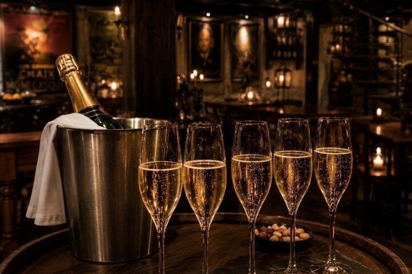A bottle of champagne in a bucket, five glasses of champagne, and a bowl of cherries on a wooden table in a dimly lit bar. 3