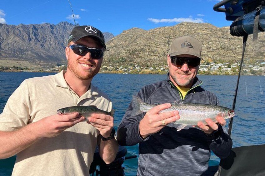 Two people holding fish on a boat with mountains in the background. 2