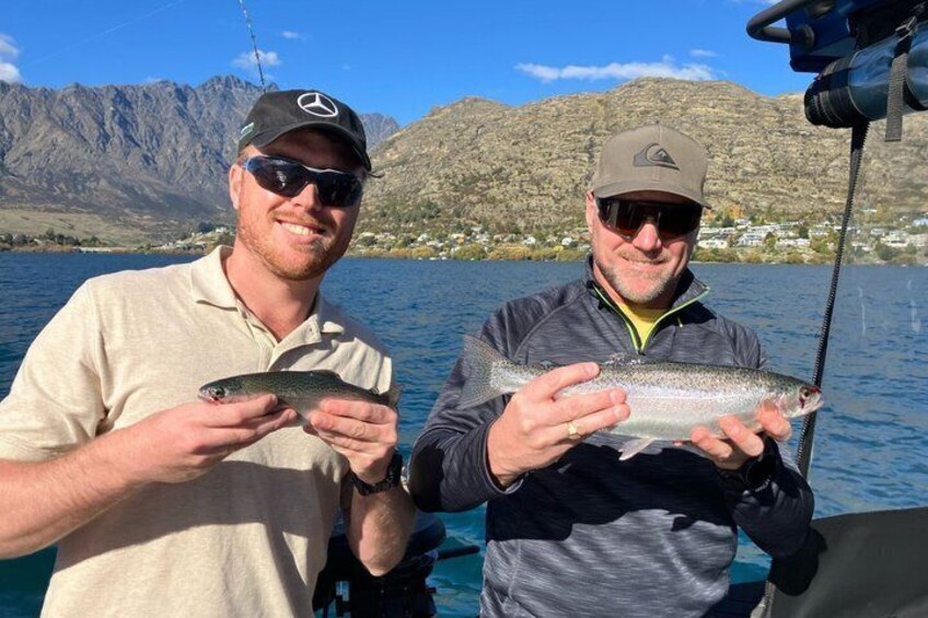 Two people holding fish on a boat with mountains in the background. 2