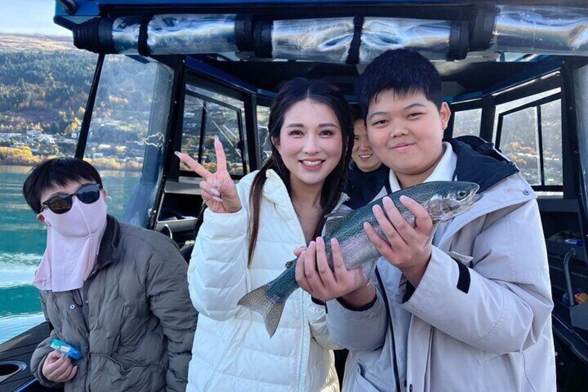 Three people on a boat holding fish, with a lake and hills in the background. 4
