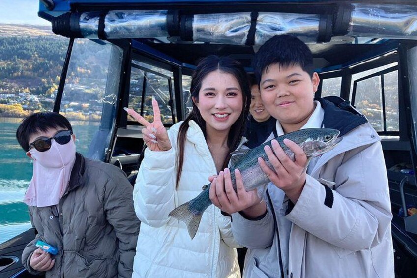 Three people on a boat holding fish, with a lake and hills in the background. 4