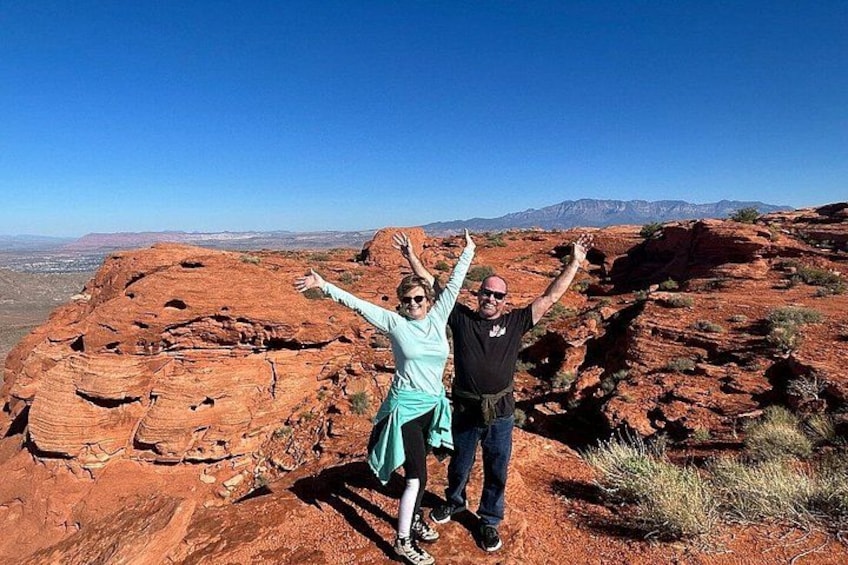 Two people standing on a red rock formation with arms raised, a clear blue sky, and distant mountains. 3