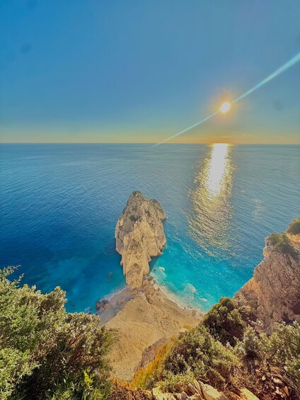 A coastal landscape with a prominent rock formation, clear blue sea, and a setting sun. 1