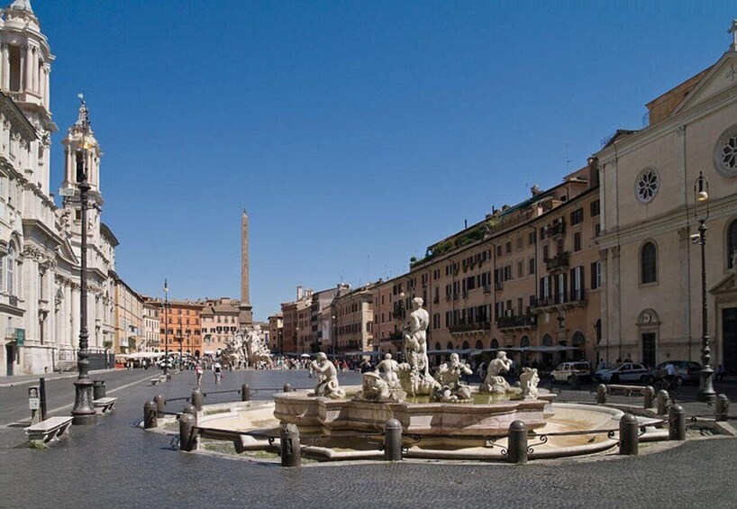 A historic square with a central fountain, classical architecture, and a prominent obelisk in the background. 3