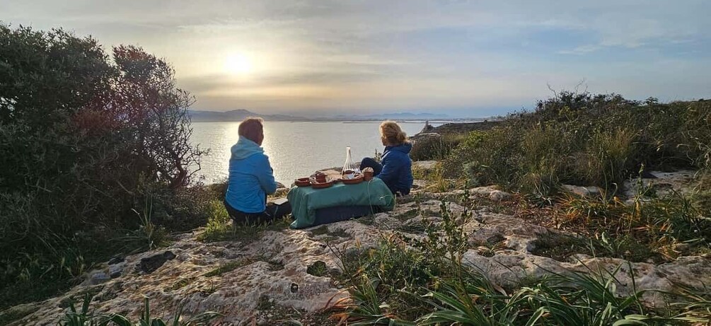 Two people sitting on a rocky outcrop overlooking a body of water with a sunset in the background. 3