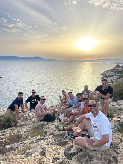 A group of people sitting on rocks by a body of water with a sunset in the background. 4