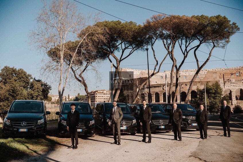 Men in suits standing in front of parked cars, with trees and a building in the background. 1