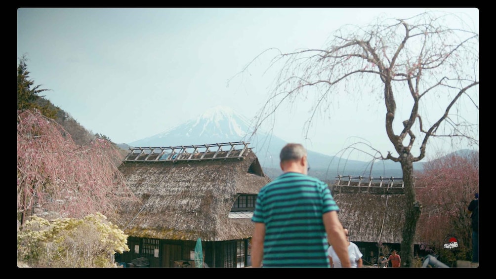A man walking towards a traditional thatched-roof house with a mountain in the background. 1