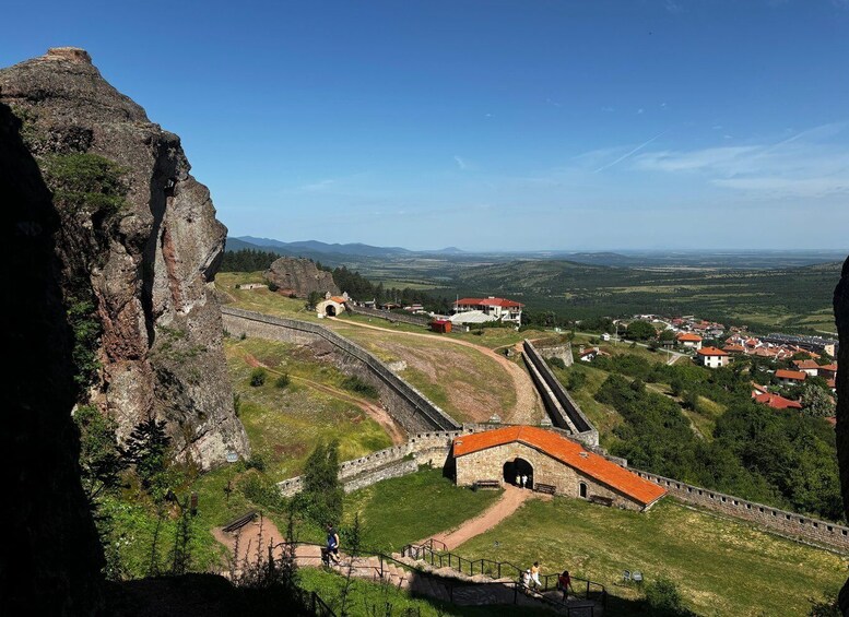A historic fortification with a red-roofed building, a stone wall, and a view of a town in the valley below. 5