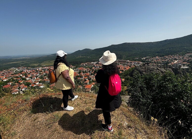 Two people standing on a hill overlooking a town with red-roofed houses and green hills in the background. 2